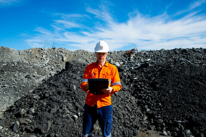 News 14 A person in an orange safety jacket and white hard hat stands in front of a heap of rocks and soil, holding a tablet that details the history of XRF. The sky is blue with wispy clouds.