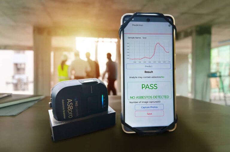 A handheld asbestos detector and a smartphone displaying a PASS result for an asbestos test sit on a table at a construction site, with three blurred people in safety gear ensuring asbestos safety in the background.