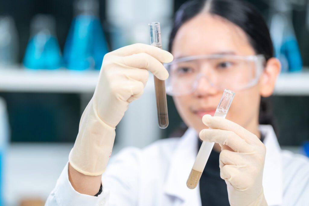 News 18 A scientist, equipped with safety goggles and gloves, conducts a lab analysis by comparing two test tubes—one containing a brown liquid and the other a pale one—set against a backdrop of blurred blue bottles in the laboratory.