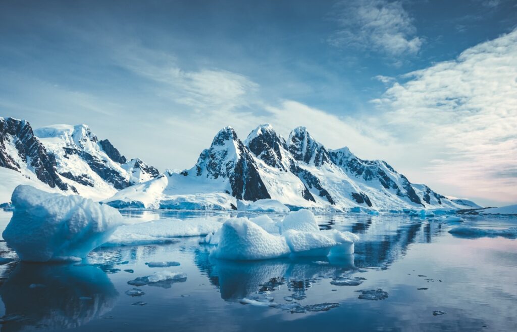 News 2 Snow-covered Antarctic mountains rise behind floating icebergs on calm blue water under a partly cloudy sky.