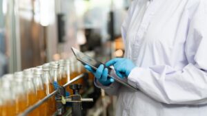 Person in a white lab coat and blue gloves using a tablet to inspect bottles of orange liquid on a beverage production line.